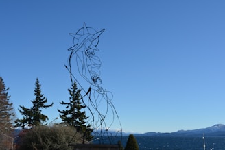 A metal wire sculpture depicting a stylized figure with an outstretched arm holding a child, set against a backdrop of blue sky and mountains in the distance. There are trees in the foreground and a large body of water behind the sculpture.