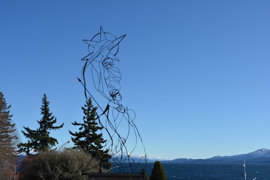 A metal wire sculpture depicting a stylized figure with an outstretched arm holding a child, set against a backdrop of blue sky and mountains in the distance. There are trees in the foreground and a large body of water behind the sculpture.