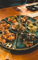A round ceramic platter with multiple sections filled with various sushi and sashimi dishes, including sliced fish, garnished rolls, and vegetables like cucumber slices. The setting appears to be a cozy restaurant with wooden tables.