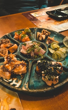 A round ceramic platter with multiple sections filled with various sushi and sashimi dishes, including sliced fish, garnished rolls, and vegetables like cucumber slices. The setting appears to be a cozy restaurant with wooden tables.