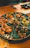 A round ceramic platter with multiple sections filled with various sushi and sashimi dishes, including sliced fish, garnished rolls, and vegetables like cucumber slices. The setting appears to be a cozy restaurant with wooden tables.