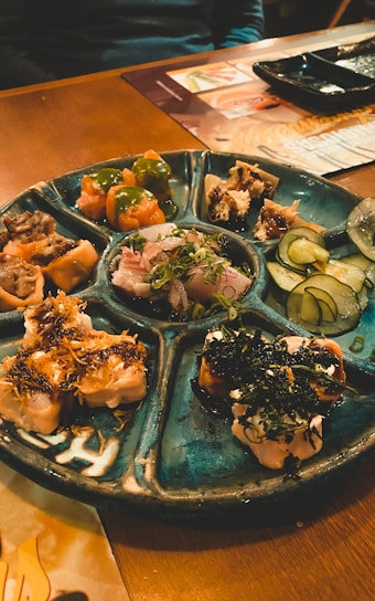 A round ceramic platter with multiple sections filled with various sushi and sashimi dishes, including sliced fish, garnished rolls, and vegetables like cucumber slices. The setting appears to be a cozy restaurant with wooden tables.