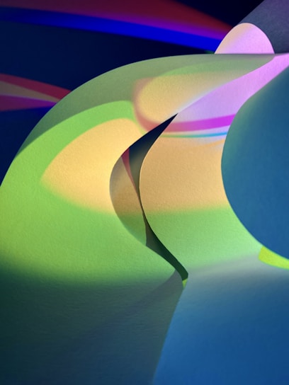 Abstract shapes and forms created by layered sheets of colored paper illuminated by dramatic lighting. The interplay of light and shadow creates a dynamic and vibrant composition with smooth, curved lines.