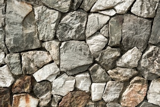 A close-up view of a stone wall made from variously-sized, rough-textured rocks that fit together without visible mortar. The stones range in color from gray to brown, exhibiting natural imperfections and irregular shapes. The overall structure appears solid and rustic.