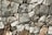A close-up view of a stone wall made from variously-sized, rough-textured rocks that fit together without visible mortar. The stones range in color from gray to brown, exhibiting natural imperfections and irregular shapes. The overall structure appears solid and rustic.