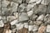 A close-up view of a stone wall made from variously-sized, rough-textured rocks that fit together without visible mortar. The stones range in color from gray to brown, exhibiting natural imperfections and irregular shapes. The overall structure appears solid and rustic.