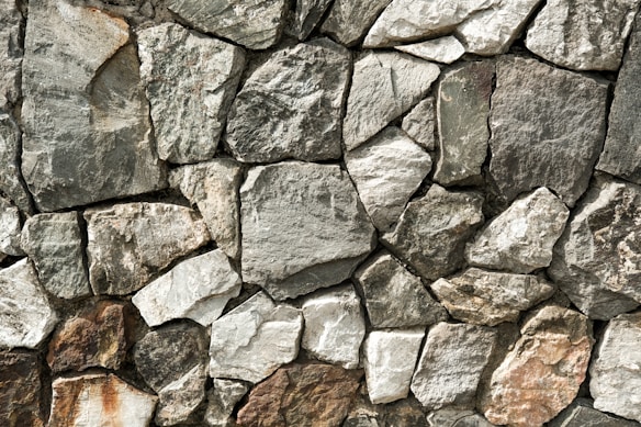 A close-up view of a stone wall made from variously-sized, rough-textured rocks that fit together without visible mortar. The stones range in color from gray to brown, exhibiting natural imperfections and irregular shapes. The overall structure appears solid and rustic.