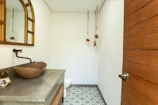 A small, modern bathroom features a wooden countertop with a stone vessel sink and a wall-mounted faucet. A large mirror with a curved top hangs above the sink, and three small potted plants are suspended from the ceiling. The floor is decorated with patterned tiles, and a wooden door with a metal handle is visible.