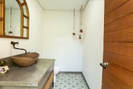 A small, modern bathroom features a wooden countertop with a stone vessel sink and a wall-mounted faucet. A large mirror with a curved top hangs above the sink, and three small potted plants are suspended from the ceiling. The floor is decorated with patterned tiles, and a wooden door with a metal handle is visible.