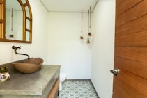 A small, modern bathroom features a wooden countertop with a stone vessel sink and a wall-mounted faucet. A large mirror with a curved top hangs above the sink, and three small potted plants are suspended from the ceiling. The floor is decorated with patterned tiles, and a wooden door with a metal handle is visible.