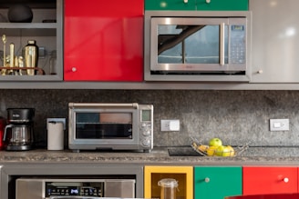 A modern kitchen counter features a variety of appliances and decor. There are bright, multicolored cabinets with vibrant red, yellow, and green shades. A microwave and a toaster oven are placed prominently on the countertop. Below, there is a coffee maker and paper towel holder. A bowl of apples and bananas adds a fresh, lively touch. Metallic elements, like a shaker and other kitchen tools, are visible on the upper shelf.