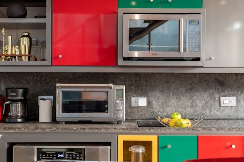 A modern kitchen counter features a variety of appliances and decor. There are bright, multicolored cabinets with vibrant red, yellow, and green shades. A microwave and a toaster oven are placed prominently on the countertop. Below, there is a coffee maker and paper towel holder. A bowl of apples and bananas adds a fresh, lively touch. Metallic elements, like a shaker and other kitchen tools, are visible on the upper shelf.