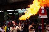 A performer is entertaining a crowd by breathing fire, creating a large flame. The setting appears to be an outdoor celebration with a red banner wishing a Happy Chinese New Year. Numerous people, including children, are watching the performance, some smiling and appearing intrigued.