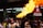 A performer is entertaining a crowd by breathing fire, creating a large flame. The setting appears to be an outdoor celebration with a red banner wishing a Happy Chinese New Year. Numerous people, including children, are watching the performance, some smiling and appearing intrigued.