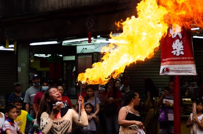 A performer is entertaining a crowd by breathing fire, creating a large flame. The setting appears to be an outdoor celebration with a red banner wishing a Happy Chinese New Year. Numerous people, including children, are watching the performance, some smiling and appearing intrigued.