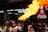 A performer is entertaining a crowd by breathing fire, creating a large flame. The setting appears to be an outdoor celebration with a red banner wishing a Happy Chinese New Year. Numerous people, including children, are watching the performance, some smiling and appearing intrigued.