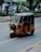 A yellow and black auto-rickshaw is moving along an urban street. The background includes parked cars, people walking, greenery, and a large tree on the right. The road is lined with paving stones and surrounded by small bushes.