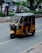 A yellow and black auto-rickshaw is moving along an urban street. The background includes parked cars, people walking, greenery, and a large tree on the right. The road is lined with paving stones and surrounded by small bushes.