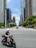 Man riding a sleek scooter on a sunny city street with skyscrapers and park greenery visible.