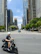 Man riding a sleek scooter on a sunny city street with skyscrapers and park greenery visible.