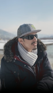 A person wearing a checkered brim cap and sunglasses is dressed warmly with a thick coat and knitted scarf. The background shows a snowy landscape with distant mountains, and the lighting suggests a clear, cold day.