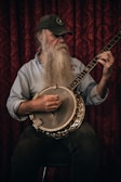A bearded man wearing a baseball cap and a denim shirt is playing a banjo. The background consists of a richly patterned red curtain. The man is focused and appears to be deeply engaged in playing the instrument.