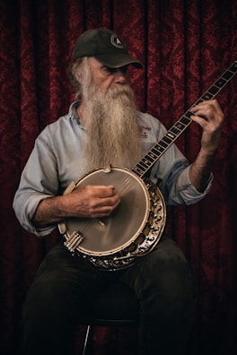 A bearded man wearing a baseball cap and a denim shirt is playing a banjo. The background consists of a richly patterned red curtain. The man is focused and appears to be deeply engaged in playing the instrument.