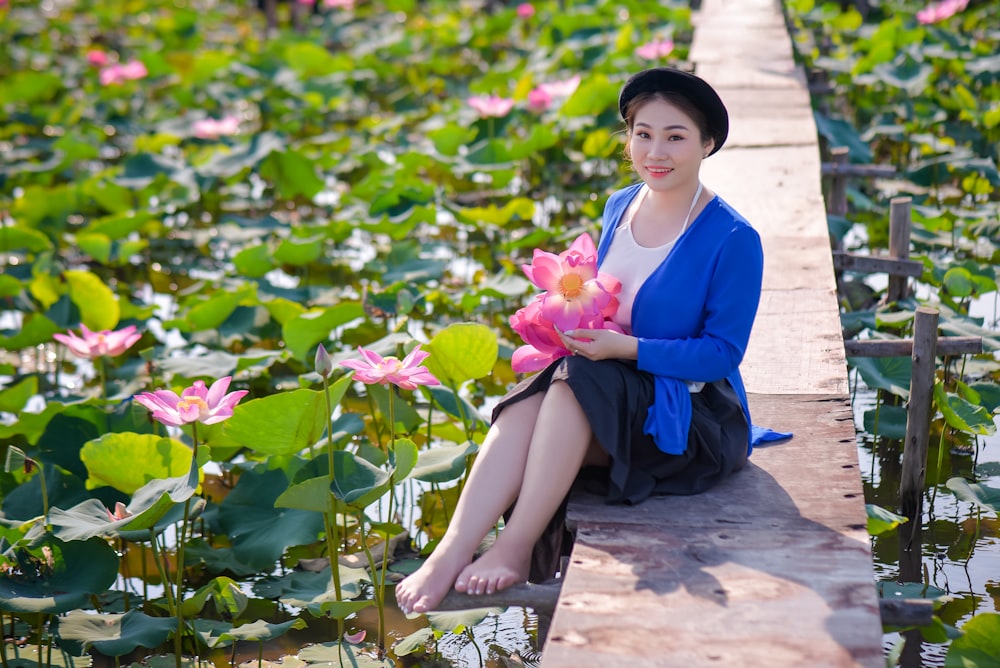 woman in blue long sleeve shirt sitting on brown wooden bench holding pink flower during daytime