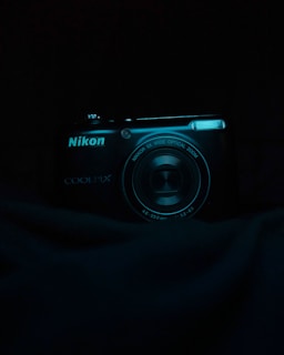 A digital camera with the brand Nikon and model Coolpix is partially illuminated against a dark background. The camera lens and branding are clearly visible, with a slight reflection on the lens surface.