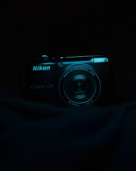 A digital camera with the brand Nikon and model Coolpix is partially illuminated against a dark background. The camera lens and branding are clearly visible, with a slight reflection on the lens surface.
