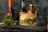 A sesame seed burger with a beef patty, lettuce, tomato, and cheese is placed on a dark wooden board surrounded by fresh lettuce leaves. In the background, there are blurred bottles and jars, possibly condiments, adding to the culinary setting.