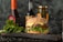 A sesame seed burger with a beef patty, lettuce, tomato, and cheese is placed on a dark wooden board surrounded by fresh lettuce leaves. In the background, there are blurred bottles and jars, possibly condiments, adding to the culinary setting.