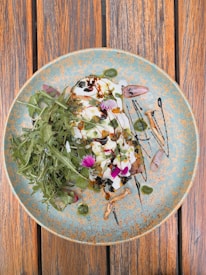 A beautifully plated dish featuring a mix of greens, garnished with vibrant edible flowers, drizzled with dark balsamic glaze. The plate is speckled and sits on a wooden table.