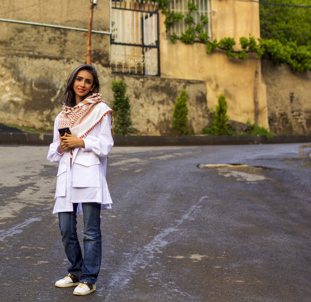 woman in white coat and blue denim jeans standing on gray asphalt road during daytime