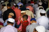 A crowded gathering of people dressed in colorful traditional attire, sitting closely together. Most individuals are wearing prayer caps, and the scene suggests a deep focus and communal activity. A young boy in a striped red shirt stands among the others.