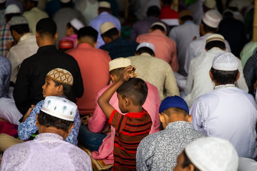 A crowded gathering of people dressed in colorful traditional attire, sitting closely together. Most individuals are wearing prayer caps, and the scene suggests a deep focus and communal activity. A young boy in a striped red shirt stands among the others.