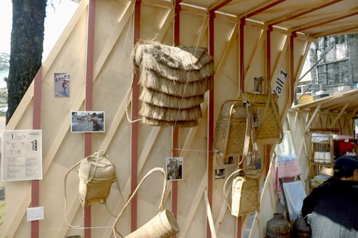 A wooden display features woven baskets and bags hanging against a wooden structure. There are framed photos and notes attached to the wall, and a person can be seen partially in the foreground. The setup appears to be part of an exhibit or market, with textiles visible in the background.