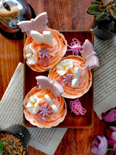 A stack of antique children's books with worn covers beside a plate of colorful cupcakes decorated with delicate frosting flowers