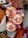 Three beautifully decorated cupcakes with intricate designs sit on a wooden surface. The frosting is tinted peach and adorned with edible flowers and butterflies. Nearby are scattered pages of a book, a decorative bell, and a small potted plant.