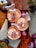 Three beautifully decorated cupcakes with intricate designs sit on a wooden surface. The frosting is tinted peach and adorned with edible flowers and butterflies. Nearby are scattered pages of a book, a decorative bell, and a small potted plant.