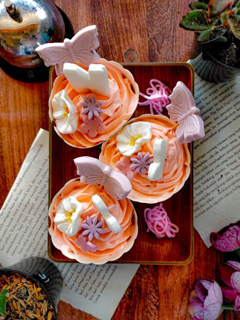 A stack of antique children's books with worn covers beside a plate of colorful cupcakes decorated with delicate frosting flowers