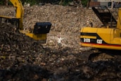 A large landfill site filled with heaps of garbage and debris. Two excavators are working amidst the piles, with one person standing between them. The scene is dominated by the dark, earthy tones of the waste, with a backdrop of trees and structures.