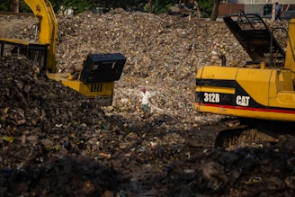A large landfill site filled with heaps of garbage and debris. Two excavators are working amidst the piles, with one person standing between them. The scene is dominated by the dark, earthy tones of the waste, with a backdrop of trees and structures.