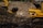 A large landfill site filled with heaps of garbage and debris. Two excavators are working amidst the piles, with one person standing between them. The scene is dominated by the dark, earthy tones of the waste, with a backdrop of trees and structures.