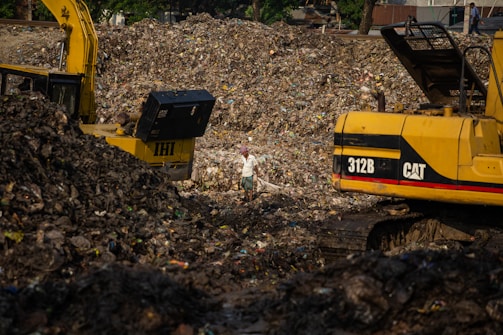 A large landfill site filled with heaps of garbage and debris. Two excavators are working amidst the piles, with one person standing between them. The scene is dominated by the dark, earthy tones of the waste, with a backdrop of trees and structures.