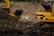 A large landfill site filled with heaps of garbage and debris. Two excavators are working amidst the piles, with one person standing between them. The scene is dominated by the dark, earthy tones of the waste, with a backdrop of trees and structures.