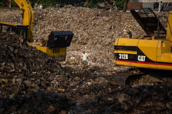 A large landfill site filled with heaps of garbage and debris. Two excavators are working amidst the piles, with one person standing between them. The scene is dominated by the dark, earthy tones of the waste, with a backdrop of trees and structures.