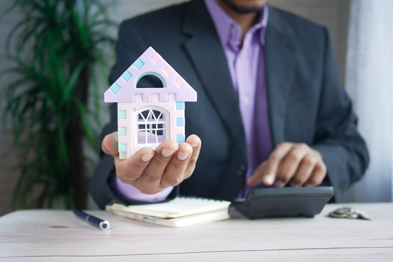 A person in a suit holds a colorful model house while using a calculator at a desk.