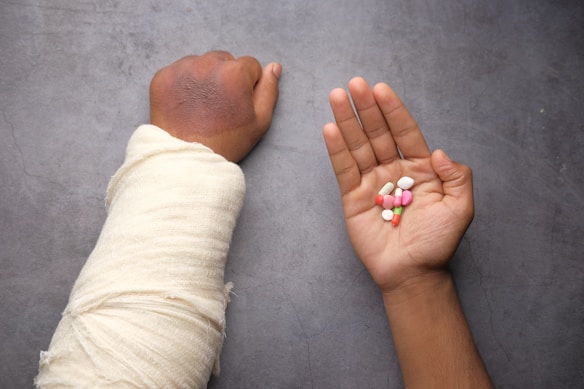 A person's arm is heavily bandaged, indicating an injury. The other hand holds a variety of colorful pills and tablets on the palm. The background is a plain gray surface.