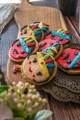 Colorful cookies decorated with icing to resemble faces with distinctive features, such as red lips, black eyes, and multicolored headdresses. The cookies are placed on a wooden cutting board, with intricate doily details beside them, creating a playful and artistic presentation.